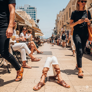 A vibrant, sun-drenched street scene in Tel Aviv, Israel. The focus is on a diverse group of people wearing unique, handcrafted leather sandals.